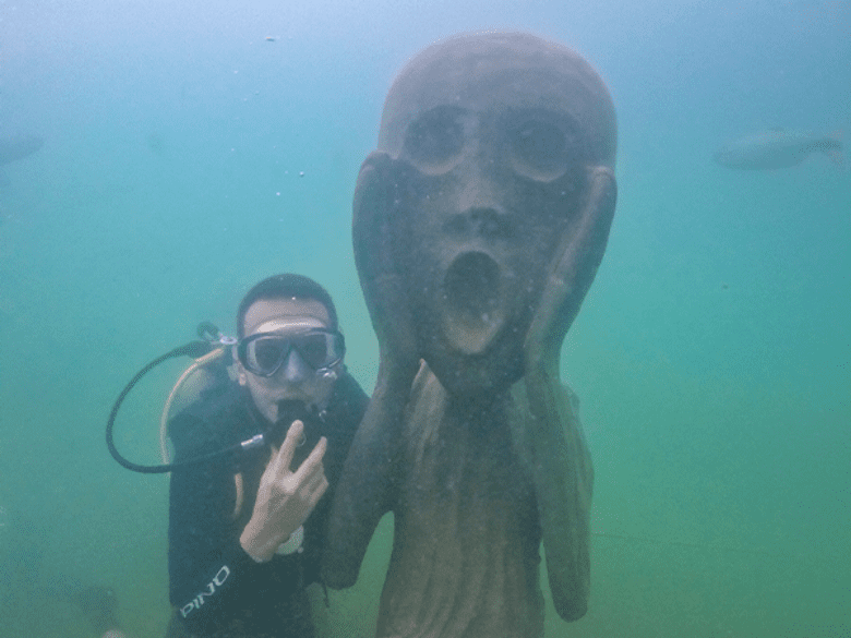 Pessoa debaixo d’água com máscara e aparelho de respiração por meio de cilindro para mergulho ao lado de uma escultura feita com resíduos naturais do fundo do rio da Nascente Azul - experiências incluídas no combo Museu Subaquático e Balneário - descontos em Bonito Ms
