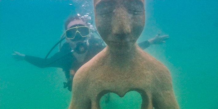 Pessoa em mergulho com cilindro atrás de uma estátua feita com recursos naturais encontrados no fundo do rio em Museu Subaquático na Nascente Azul