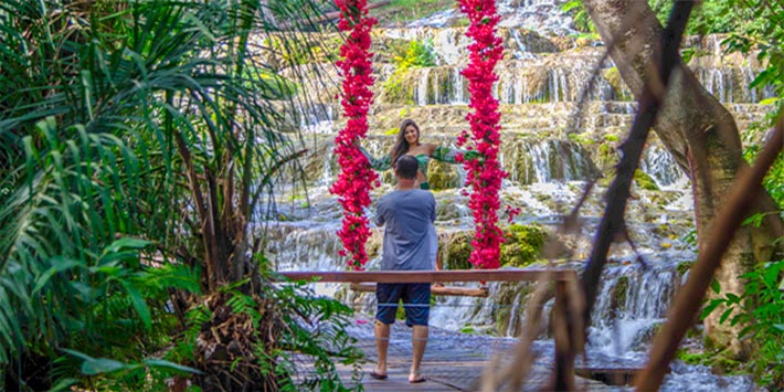 Casal tirando foto no balanço florido de frente para cachoeira de tufas calcárias, repleta de natureza ao redor - Balneário Nascente Azul