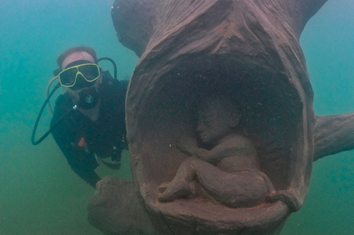 Pessoa em mergulho com cilindro passando por escultura submersa em museu subaquático - passeios com crianças em Bonito MS