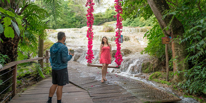 Casal contemplando a cachoeira de tufas calcárias que deságua em forma de véu, cercada de vegetação nativa e com um balanço de flores rosas à frente - uma das paradas das trilhas acessíveis dentro da Nascente Azul em Bonito MS