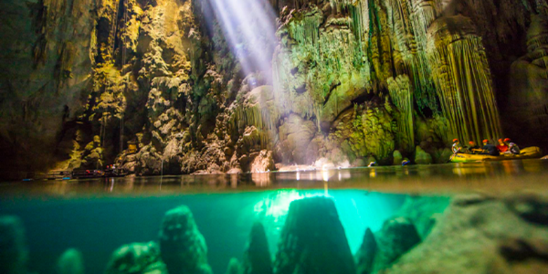 Imagem de lago na parte interna do Abismo Anhumas, com parte da fotografia mostrando as formações rochosas debaixo d’água, e a outra parte mostrando um bote com turistas navegando pelo lago, em meio a um facho de luz no meio da caverna.