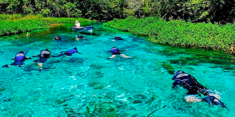 Grupo de turistas flutua sob as águas azuis e cristalinas do Rio Sucuri, com um barco ao fundo da imagem, e o rio cercado por vegetação, em dia ensolarado.