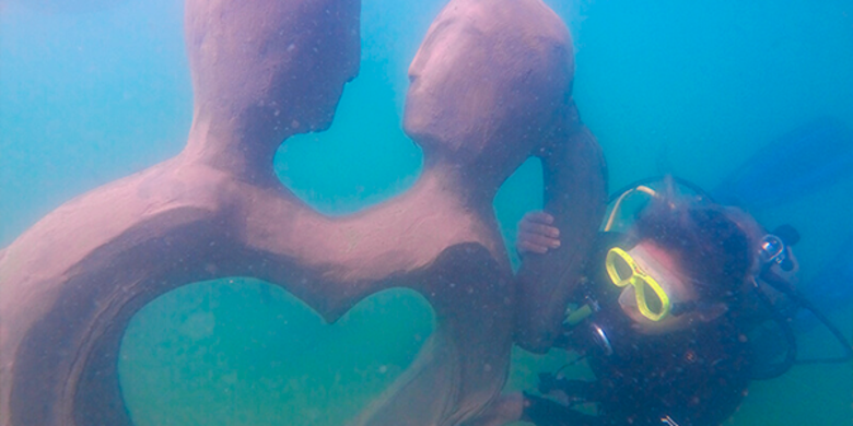 Mergulhadora posa ao lado de escultura com um casal se encarando, em formato de coração, no fundo do rio. 