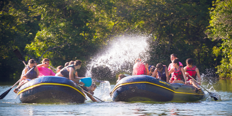 Dois botes levam grupo de turistas, que jogam baldes de água uns nos outros, pelo rio em dia ensolarado.